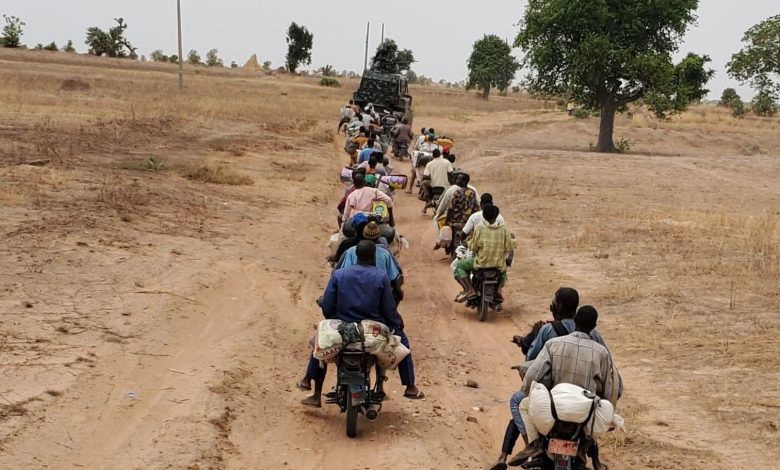 People on motorcycles follow a military vehicle on a dirt road through a dry landscape with sparse trees.