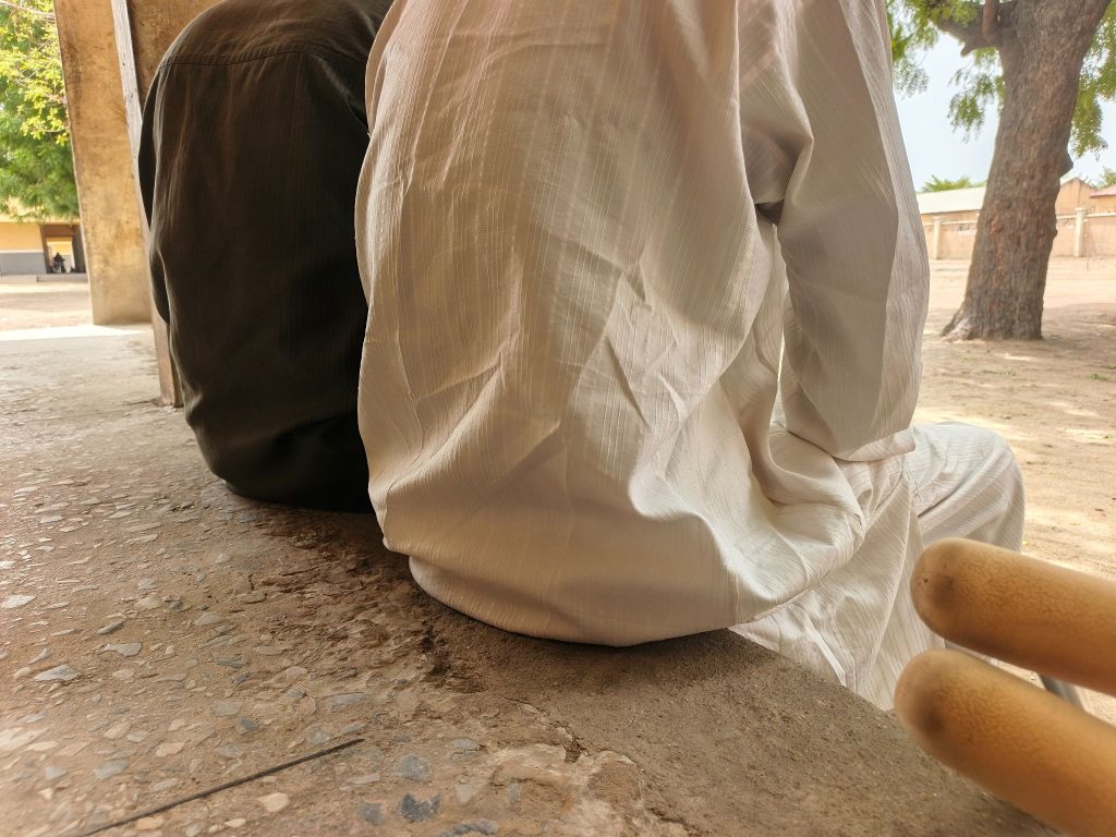 Two people sitting side by side on a ledge, one in brown and one in white. There's a tree in the background and textured ground.