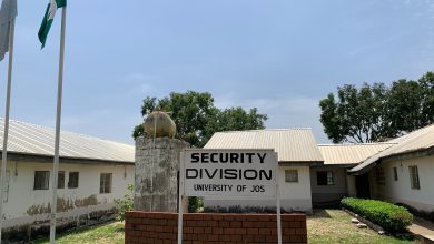 Security Division sign at University of Jos, flanked by flagpoles, with a white building and trees in the background.