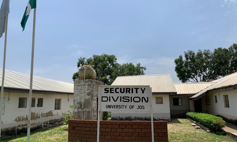 Security Division sign at University of Jos, flanked by flagpoles, with a white building and trees in the background.
