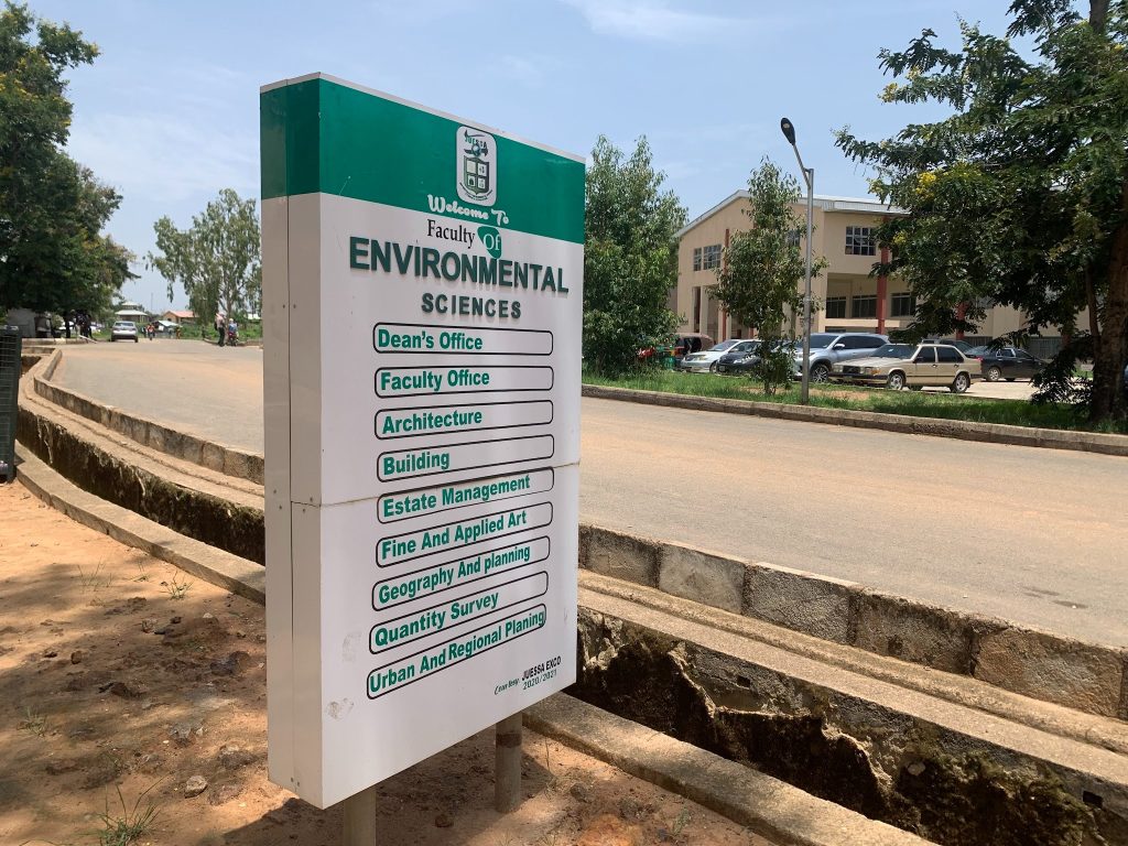 Sign for the Faculty of Environmental Sciences by a road, listing offices like Architecture and Geography. Trees and a building in the background.