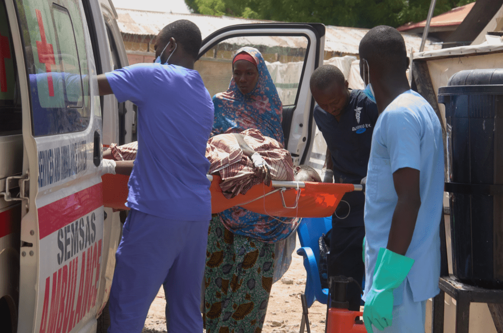 Medical personnel load a patient onto an ambulance in a rural area, while a woman stands nearby.