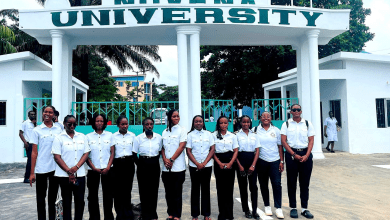 A group of people in uniform standing in front of Novena University entrance with a white gate and trees in the background.
