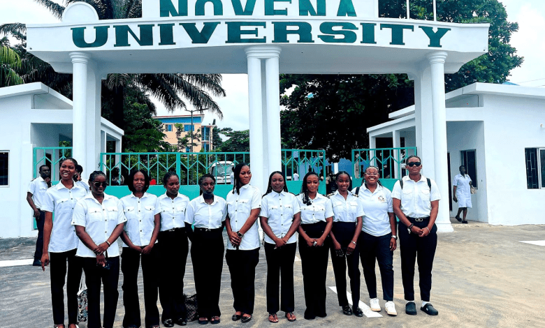 A group of people in uniform standing in front of Novena University entrance with a white gate and trees in the background.