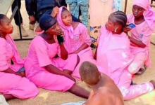 A group of people, mostly in pink attire, sit on the ground, engaging in conversation with others standing around them.