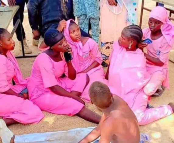 A group of people, mostly in pink attire, sit on the ground, engaging in conversation with others standing around them.