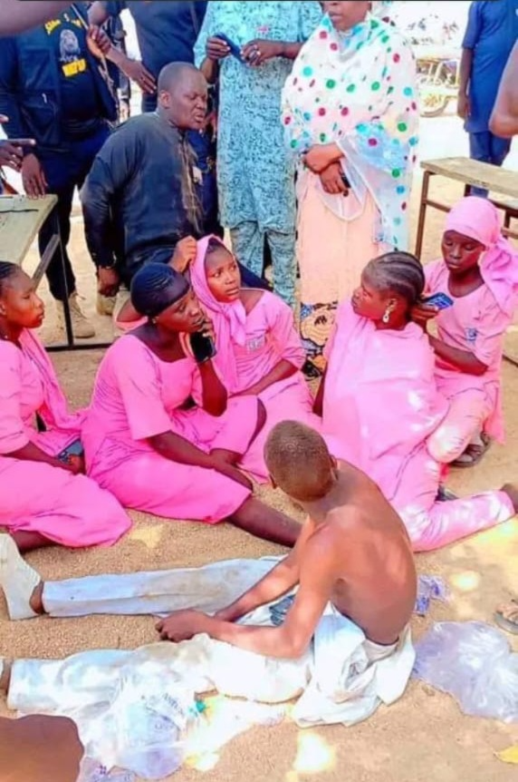 A group of people, mostly in pink attire, sit on the ground, engaging in conversation with others standing around them.