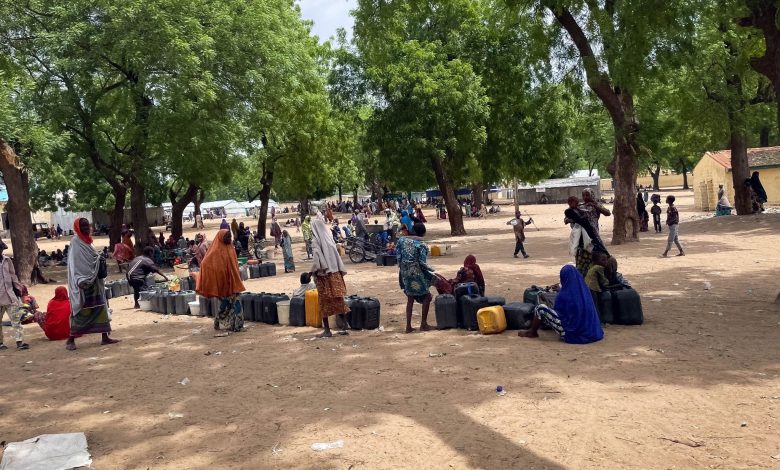 People gather under trees, with containers, in a sandy open area, while others are in the background near buildings.