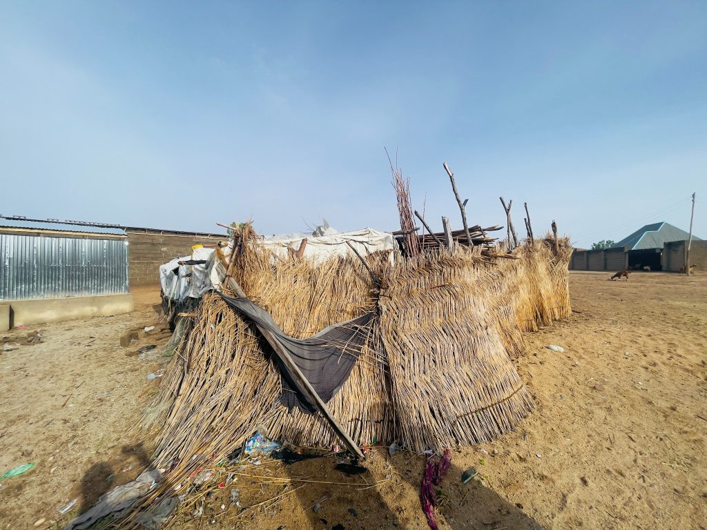 Tattered hut made of sticks and tarps in a dry, barren area, with other structures visible in the background under clear blue sky.