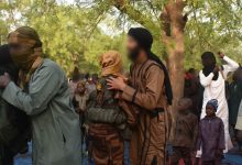 A group of people in the outdoors, some wearing camouflage and face coverings, others in traditional attire. Trees are in the background.