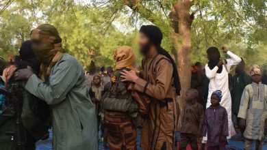 A group of people in the outdoors, some wearing camouflage and face coverings, others in traditional attire. Trees are in the background.