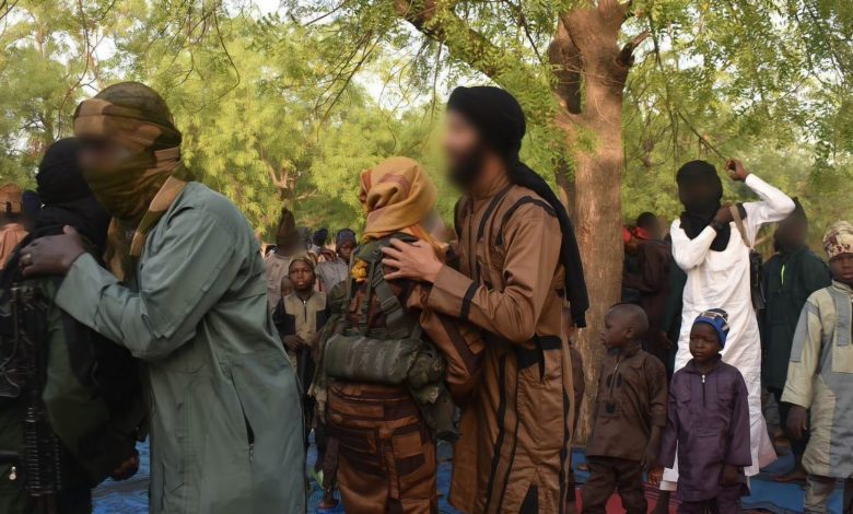 A group of people in the outdoors, some wearing camouflage and face coverings, others in traditional attire. Trees are in the background.