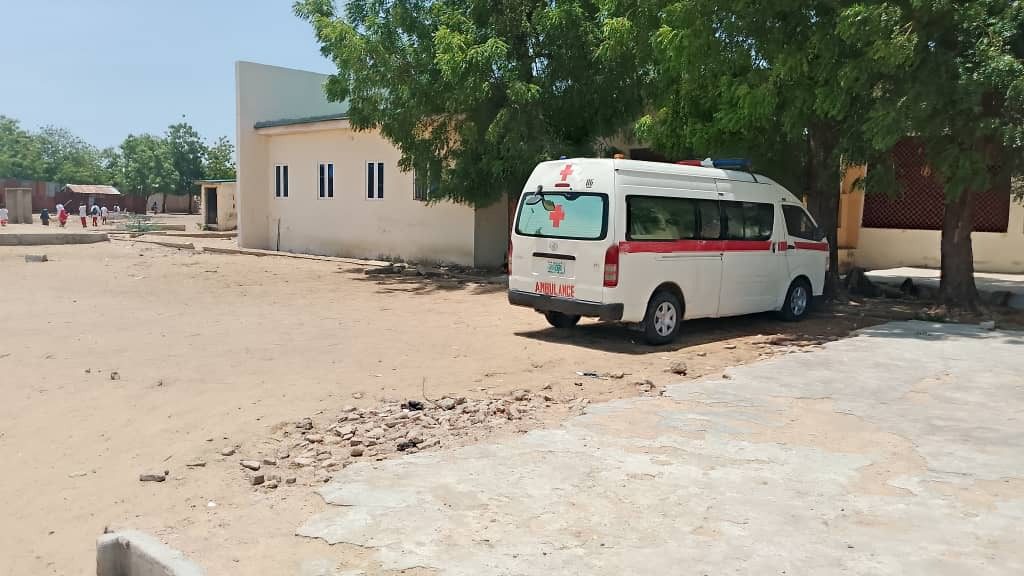 An ambulance is parked under a tree near a light-colored building in a sandy area on a sunny day.