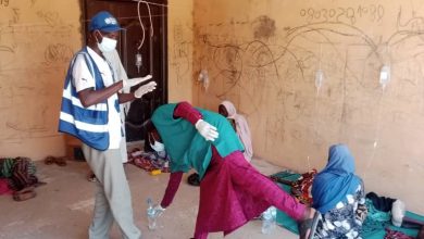Healthcare workers assist individuals in a room with IV drips, graffiti on walls, and people sitting or lying on mats.