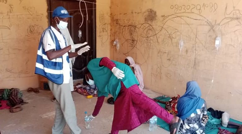 Healthcare workers assist individuals in a room with IV drips, graffiti on walls, and people sitting or lying on mats.