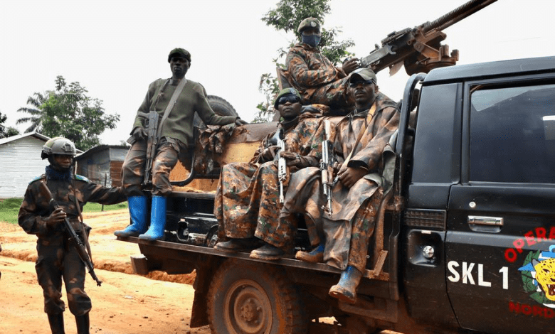 Soldiers in camo sit on a truck mounted with a gun, while another stands nearby with a rifle. Trees and structures in the background.