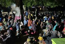 A large group of people, including children, sitting under a tree at night. Many are wearing colorful clothing and head coverings.