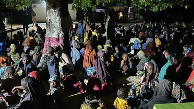 A large group of people, including children, sitting under a tree at night. Many are wearing colorful clothing and head coverings.