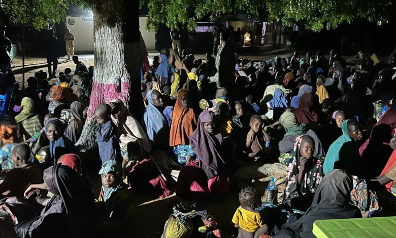 A large group of people, including children, sitting under a tree at night. Many are wearing colorful clothing and head coverings.