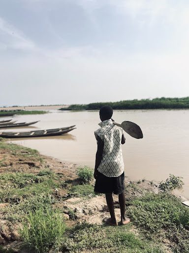 A person holding a paddle stands by a riverbank, looking at the water. Nearby, some wooden boats are docked.