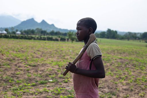 Young person holding a tool, standing in a field with distant mountains in the background.