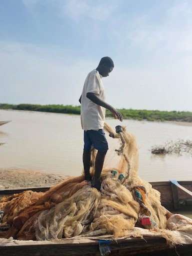 A person stands on a small boat, sorting fishing nets by a calm river or lake under a blue sky.