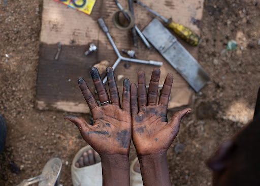 Greasy hands held up, with scattered tools and a dirt ground beneath.