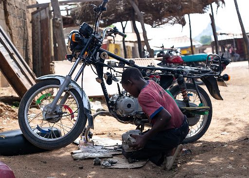 Person repairing a motorcycle outdoors, surrounded by tools and parts, under a thatched shelter.