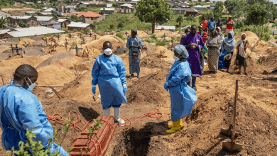 Healthcare workers in protective gear bury a coffin as onlookers stand by, in a hilly village cemetery under a blue sky.