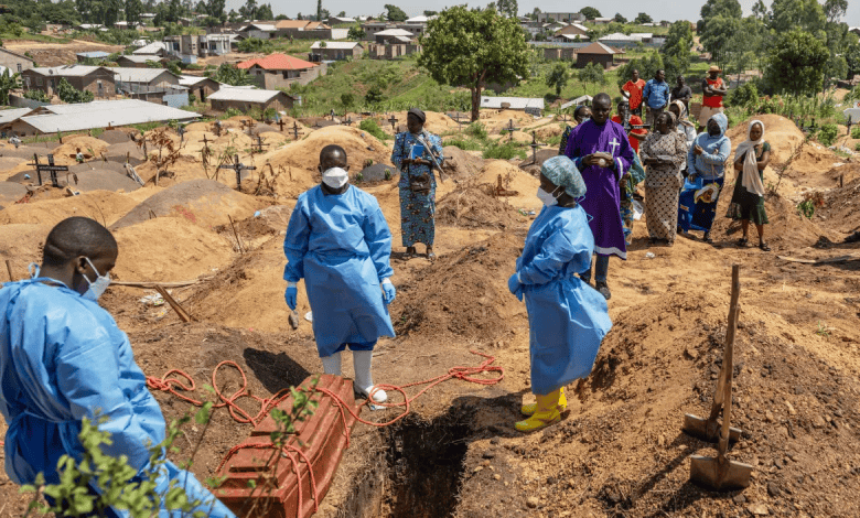 Healthcare workers in protective gear bury a coffin as onlookers stand by, in a hilly village cemetery under a blue sky.
