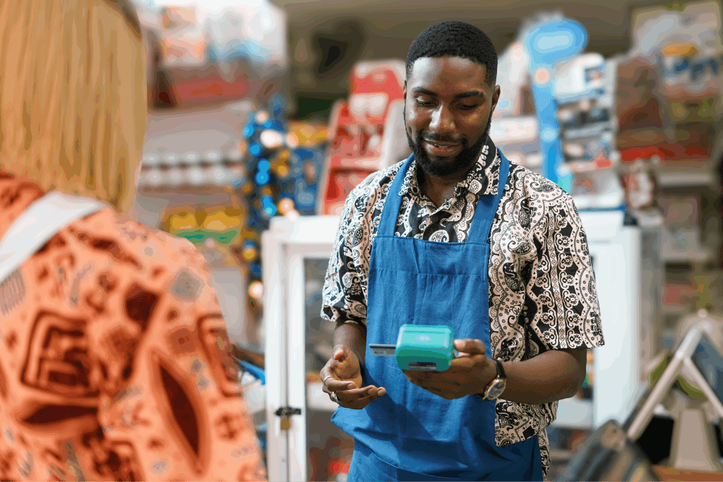 Store clerk in a blue apron processing a payment with a card reader while interacting with a customer at the counter.