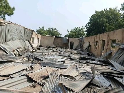Debris of a collapsed building with corrugated metal sheets scattered on the ground under a clear sky. Trees are visible in the background.