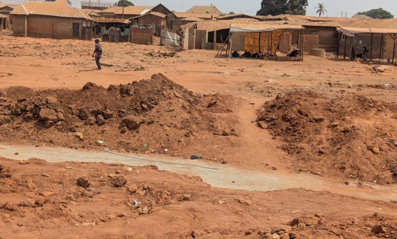 A lone person walks across a dry, rugged, reddish-brown landscape with a background of rustic buildings under a clear sky.