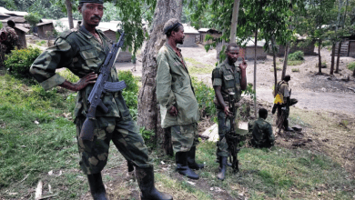 Soldiers in camouflage stand with rifles under trees in a rural setting with wooden buildings in the background.