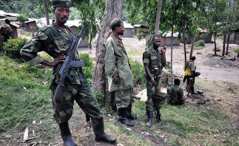 Soldiers in camouflage stand with rifles under trees in a rural setting with wooden buildings in the background.