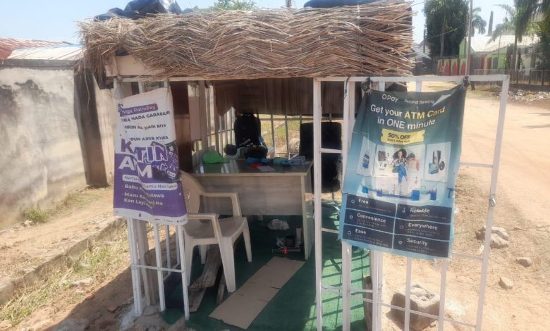Small outdoor booth with straw roof, advertising ATM and banking services. Two banners hang at the entrance, with chairs and a table inside.