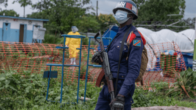 A masked peacekeeper stands with a rifle in a fenced area, while a person in protective gear walks in the background.