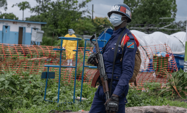 A masked peacekeeper stands with a rifle in a fenced area, while a person in protective gear walks in the background.