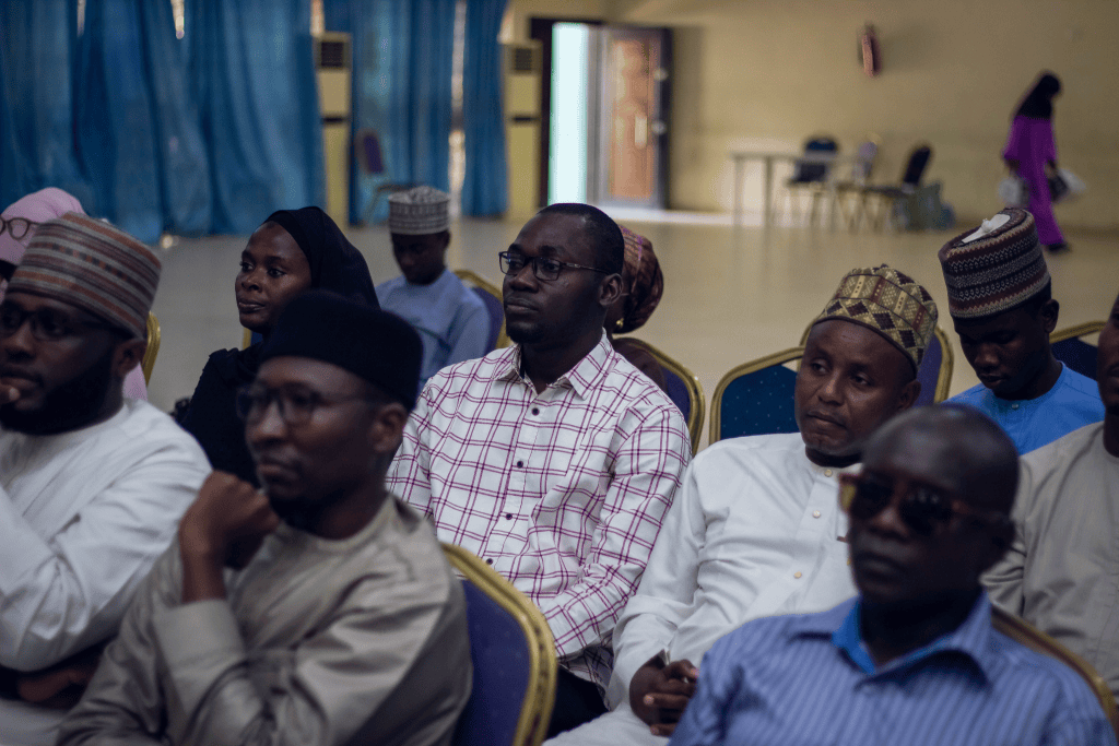 People seated attentively in a room with blue curtains and empty chairs in the background.