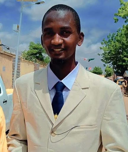 Smiling person in a light suit and tie outdoors, with trees and a bright sky in the background.