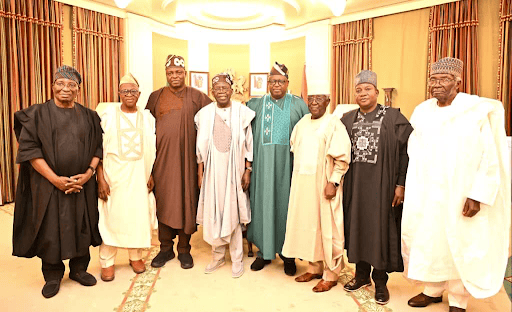 A group of eight men wearing traditional Nigerian attire stand together, posing for a photo in an elegantly decorated room.