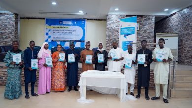 Group of people at a book launch event, each holding a copy of the book. Banners and a table with a white cloth are in the background.