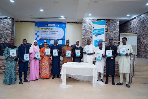 Group of people at a book launch event, each holding a copy of the book. Banners and a table with a white cloth are in the background.