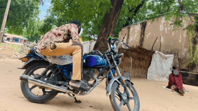 A person sits on a motorcycle parked on a sandy ground, with trees in the background and another person sitting nearby.