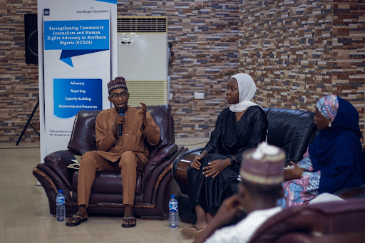 People in discussion at a community journalism and human rights event, sitting on chairs with a banner in the background.