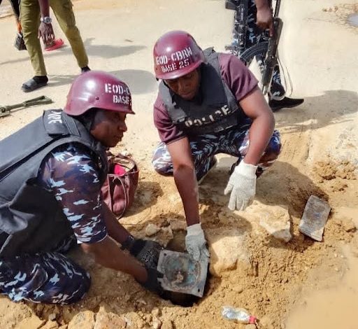 Two uniformed individuals examining an object in a small excavation, wearing protective helmets and gloves, with a third person in the background.